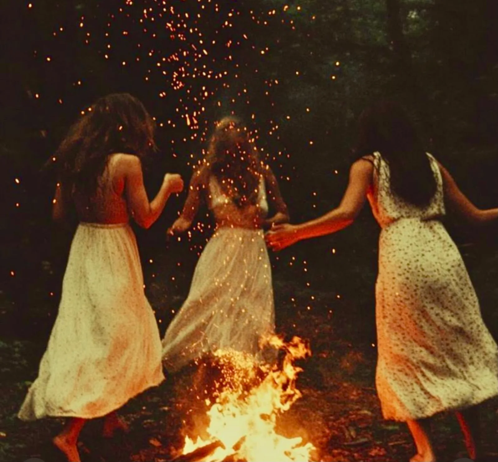 Three women in white dresses gathered around a fire at night