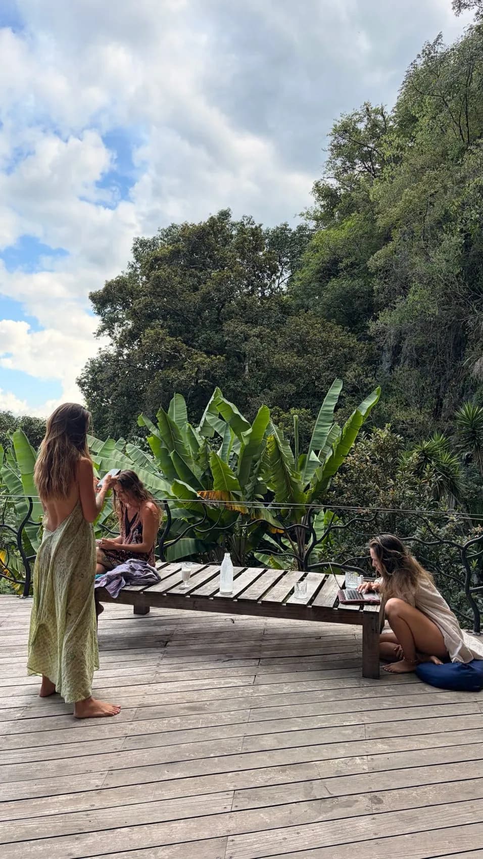 Women on the deck among banana leaves, one standing, one on her phone, one working at a laptop