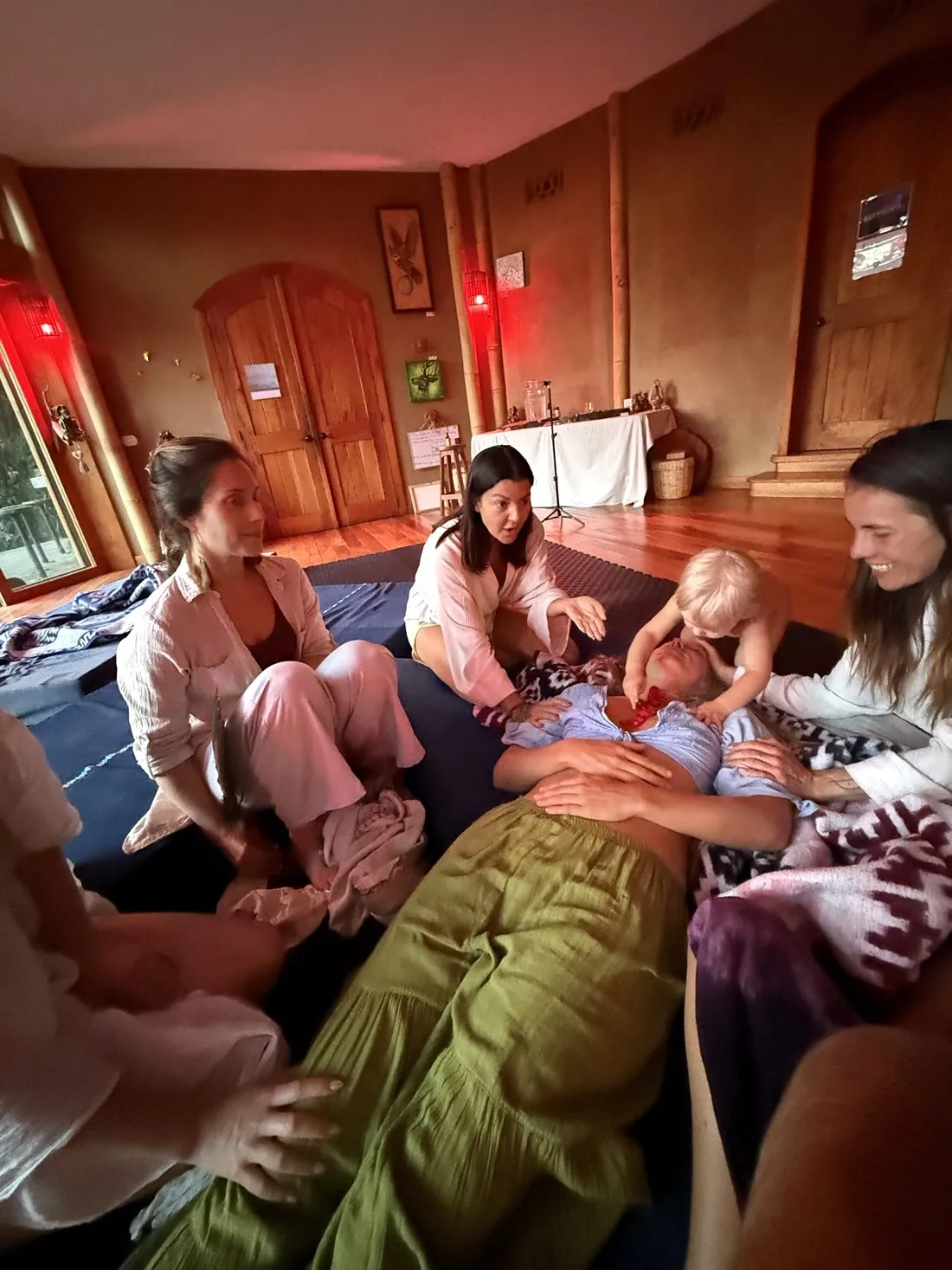 Sisters with their hands resting on a woman lying down, a toddler leaning in, in a red-lit room