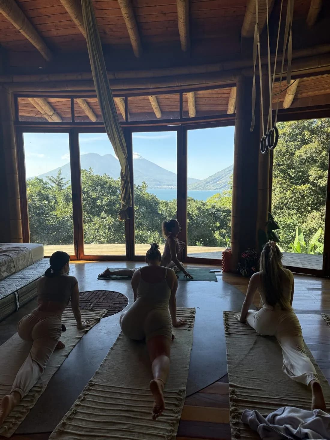 Women stretching on woven mats in the studio, looking out through tall windows to the lake and volcano