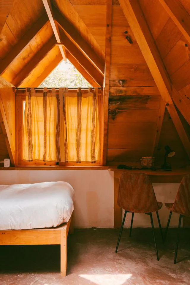 A twin bed by a pyramid window with curtains, a desk and chairs