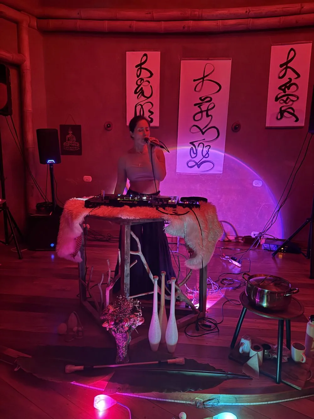 A woman singing into a microphone at a sound table in a pink and red lit dome, beneath calligraphy banners