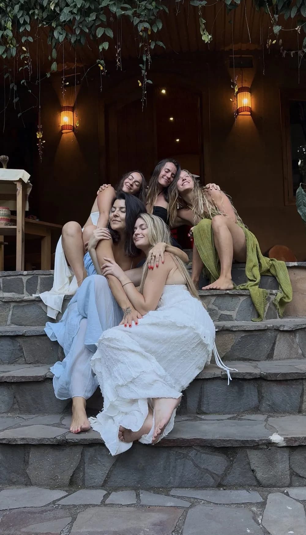 Five sisters embracing on stone steps beneath glowing lanterns, eyes closed, heads resting together