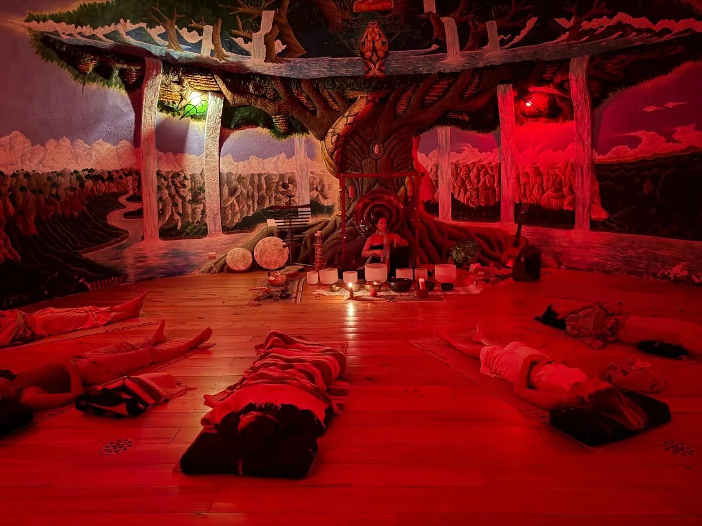 Women resting during a sound bath in a red-lit room beneath a painted tree mural